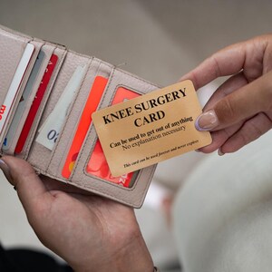 Op de afbeelding: Een beige portemonnee met diverse creditcards en een rechthoekige goudkleurige kaart met zwarte tekst: KNEE SURGERY CARD, Can be used to get out of anything, No explanation necessary, This card can be used forever and never expires.