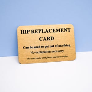 Op de afbeelding: Een rechthoekige, lichtgekleurde houten kaart met afgeronde hoeken en zwarte tekst: HIP REPLACEMENT CARD, Can be used to get out of anything, No explanation necessary, This card can be used forever and never expires.