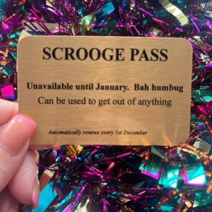 以下が含まれることがあります： カラフルなキラキラした背景に、黒い文字で「SCROOGE PASS」と書かれた金色のカード。カードには「1月まで利用不可。Bah humbug。何でも脱出できる。毎年12月1日に自動更新」と書かれています。