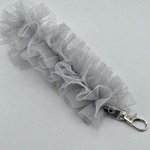 May include: A gray tulle wrist strap with a ruffled design. The strap has a silver-colored metal clasp and a small, dark gray leather accent. The tulle is layered, creating a textured, decorative look.