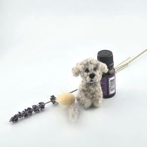 May include: A small, needle-felted gray and white dog figurine with a textured, fuzzy appearance, positioned next to a dark brown essential oil bottle with a purple label and a sprig of dried lavender and fluffy ornamental grass.