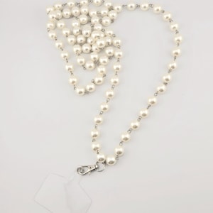 May include: A white pearl lanyard with a clear plastic badge holder. The lanyard is made of faux pearls and silver metal.