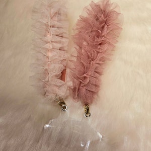 May include: Two pairs of tulle bunny ears, one in pink and one in white, with gold chain accents. The ears are on a white fuzzy surface.