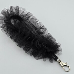 May include: Black tulle wrist strap with a silver-colored clasp. The strap is made of layered, ruffled tulle, creating a textured, decorative effect. The clasp is a standard swivel hook.