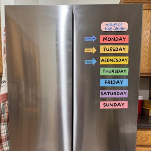 May include: A stainless steel refrigerator with colorful magnetic strips that list the days of the week. The strips are arranged vertically, with "Monday" at the top and "Sunday" at the bottom.