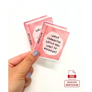 May include: Two pink watercolor-style book-shaped cards with the text "Which character would you want to befriend?" and "Book Club". The cards are held in a hand. A red PDF icon is in the bottom right.