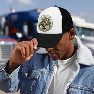 May include: Black and white trucker hat with a white front panel. The hat features a circular graphic with a vintage car and the text "Rat Rod Rampage". The hat is worn by a person.
