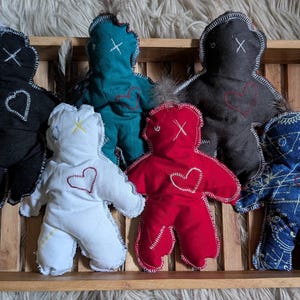 May include: A wooden tray holds six handmade cloth dolls in various colors: black, teal, white, red, dark gray, and blue. Each doll has a stitched heart and a stitched face. The dolls are arranged on a wooden tray.