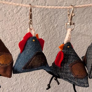 May include: Four handmade chicken-shaped keychains hanging from a rope. The keychains are made of fabric in various colors, including brown, blue denim, and black denim. Each has a red comb, orange beak, and a round patch.