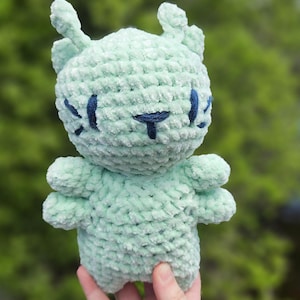 May include: A light mint green crocheted plush toy featuring a rounded head with two small antennae, four arms, and dark blue embroidered facial details including eyes and a mouth. The soft, textured yarn creates a fuzzy appearance throughout.