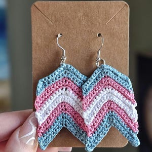May include: A pair of crocheted earrings in the colours of the transgender pride flag. The earrings are shaped like a chevron and are made with blue, pink and white yarn.