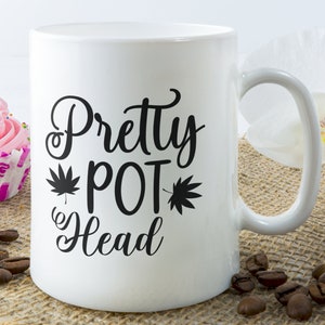 Pode incluir: Caneca de café de cerâmica branca com um design preto que diz "Pretty Pot Head" com uma folha de maconha de cada lado da palavra "Pot".