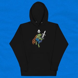 May include: Black hoodie with a graphic of a green frog wearing a pirate hat and holding a sword. The frog is wearing a blue and orange cape and has a skull and crossbones on its hat.