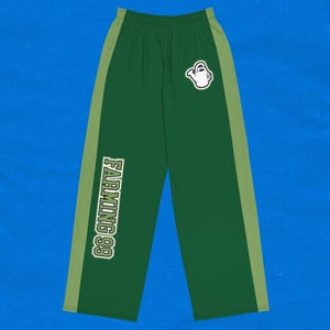 Pode incluir: Calças de moletom verdes com uma faixa lateral verde claro e o texto "FARMING 99" em branco. Um gráfico de regador branco está na perna direita. As calças estão em um fundo azul.