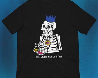 Pode incluir: Uma t-shirt preta de manga curta com um gráfico de um esqueleto branco a usar uma coroa azul e um pendente dourado. O esqueleto segura dois frascos de vidro com líquidos amarelo e rosa. O texto THE GRIND NEVER STOPS está impresso.