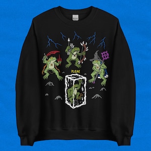 Pode incluir: Camisola preta com uma ilustração fantasiosa de rãs antropomórficas em trajes de fantasia. O design inclui rãs com armas, uma rã feiticeira e uma presa no gelo. A palavra "PLEAE" está impressa abaixo.