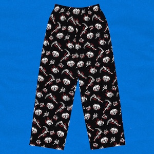 May include: Black pajama pants with a repeating pattern of white skulls, red flames, and scythes. The pants have a relaxed fit and a comfortable design, perfect for lounging or sleeping. The background is a solid blue.