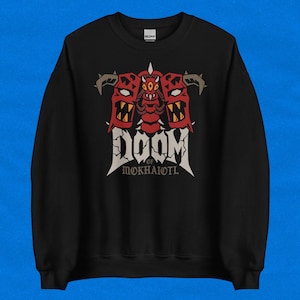 May include: Black sweatshirt with a graphic design. The design features the word "DOOM" in a stylized font, and the text "OF MOKHAIOTL". The design includes three red demon-like faces with horns and sharp teeth. The sweatshirt is made of a soft material.