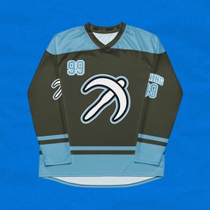 Puede incluir: Una camiseta de hockey verde oscuro con detalles en azul claro. La camiseta tiene un gráfico blanco de una letra "T" estilizada en la parte delantera. El número "99" está en el hombro izquierdo y la palabra "WING" está en el hombro derecho.