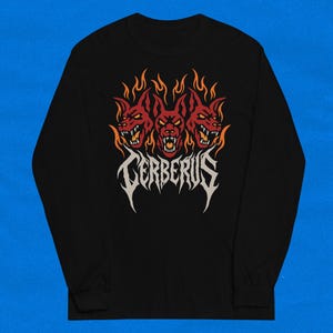 Puede incluir: Camiseta negra de manga larga con un diseño gráfico de Cerbero, el perro de tres cabezas de la mitología griega. El diseño incluye tres cabezas de perro rojas con llamas y la palabra "Cerberus" en una fuente estilizada.