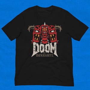 May include: Black t-shirt with a graphic design. The design features the word "DOOM" in white, and "of Mokhaiotl" in a smaller font. The graphic includes three red demon-like figures with horns and sharp teeth.