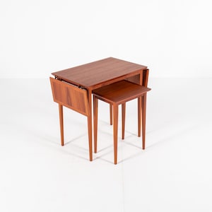 Puede incluir: Un juego de dos mesas nido con un acabado cálido de color marrón rojizo. La mesa más grande tiene una tapa rectangular con una extensión abatible, y la mesa más pequeña encaja perfectamente debajo. Ambas mesas tienen patas delgadas y cónicas.