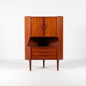 May include: A mid-century modern wooden corner cabinet with a warm brown finish. The cabinet features a drop-down desk, three drawers, and upper storage with doors. The cabinet stands on four tapered legs.