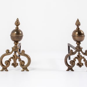 Peut inclure: Une paire de chenets de cheminée en laiton dotés de bases à volutes ornées et de fleurons sphériques surmontés d'accents en forme de flamme. Chaque chenet en métal possède une barre de support horizontale robuste s'étendant vers l'arrière.