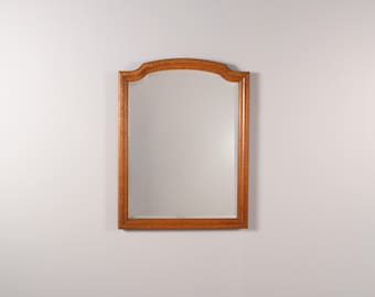 May include: A rectangular wall mirror featuring a medium-toned wooden frame with a smooth finish. The top edge of the frame is arched, while the bottom and side edges are straight. The mirror glass is clear and reflects the surrounding light.