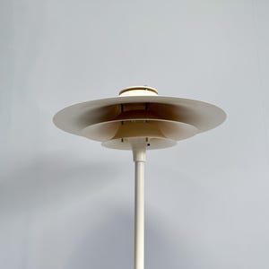 K&ouml;nnte beinhalten: Eine wei&szlig;e Stehleuchte mit mehreren Ebenen und einem kreisf&ouml;rmigen Schirmdesign. Die Lampe hat einen hohen, schlanken Stiel und mehrere sich &uuml;berlappende, scheibenf&ouml;rmige Schirme. Das Design der Lampe ist minimalistisch und modern, geeignet f&uuml;r die Inneneinrichtung.