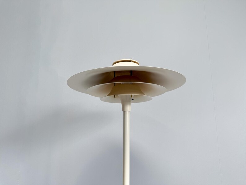 K&ouml;nnte beinhalten: Eine wei&szlig;e Stehleuchte mit mehreren Ebenen und einem kreisf&ouml;rmigen Schirmdesign. Die Lampe hat einen hohen, schlanken Stiel und mehrere sich &uuml;berlappende, scheibenf&ouml;rmige Schirme. Das Design der Lampe ist minimalistisch und modern, geeignet f&uuml;r die Inneneinrichtung.