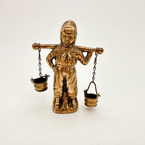 May include: A gold-colored figurine of a person carrying two buckets on a yoke. The person is wearing a hat and a long-sleeved shirt. The buckets are hanging from chains.