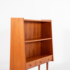 May include: A mid-century modern wooden bookcase with a warm, reddish-brown finish. The bookcase features two shelves and three small drawers with round knobs. The design includes angled sides and slender legs, showcasing a minimalist aesthetic.