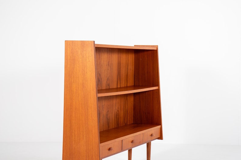 May include: A mid-century modern wooden bookcase with a warm, reddish-brown finish. The bookcase features two shelves and three small drawers with round knobs. The design includes angled sides and slender legs, showcasing a minimalist aesthetic.