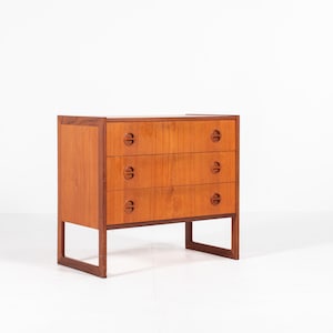 Op de afbeelding: Een houten dressoir in mid-century modern stijl met drie lades. Het dressoir is gemaakt van lichtbruin hout met een rechthoekige vorm en een eenvoudig ontwerp. Elke lade heeft een ronde handgreep. Het dressoir staat op vier poten.