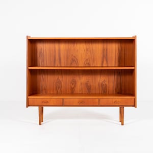 May include: A mid-century modern bookcase made of wood. The bookcase has two shelves, three drawers, and is supported by four legs. The wood has a warm, reddish-brown tone and a visible wood grain.