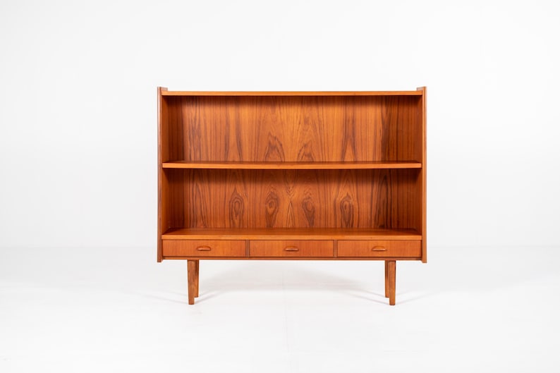 May include: A mid-century modern bookcase made of wood. The bookcase has two shelves, three drawers, and is supported by four legs. The wood has a warm, reddish-brown tone and a visible wood grain.