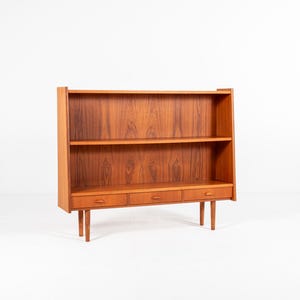 May include: A mid-century modern wooden bookcase with two shelves and three drawers. The bookcase is made of a warm-toned wood and stands on four slender legs. The design is simple and functional, with a focus on clean lines and natural materials.