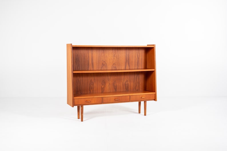 May include: A mid-century modern wooden bookcase with two shelves and three drawers. The bookcase is made of a warm-toned wood and stands on four slender legs. The design is simple and functional, with a focus on clean lines and natural materials.