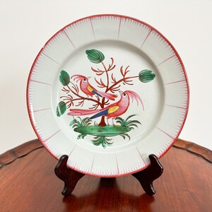 Könnte beinhalten: Ein weißer Keramikteller mit einem rosa und grünen Blumendesign mit zwei rosa Vögeln mit gelben Akzenten. Der Teller hat einen roten und weißen Rand.