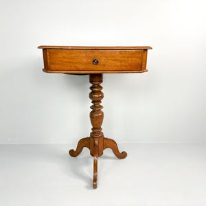 Puede incluir: Una mesa auxiliar de madera con un solo cajón y una base de pedestal torneado. La mesa tiene un acabado marrón claro y un diseño simple y elegante.