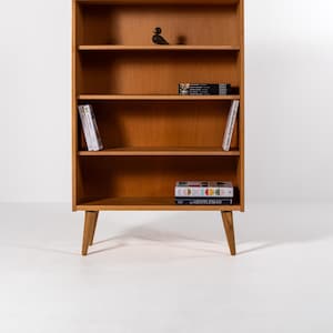 Puede incluir: Una estanter&iacute;a de madera de estilo moderno de mediados de siglo con cuatro estantes y patas anguladas. La estanter&iacute;a est&aacute; llena de libros y un peque&ntilde;o p&aacute;jaro decorativo. La madera tiene un acabado c&aacute;lido y natural.