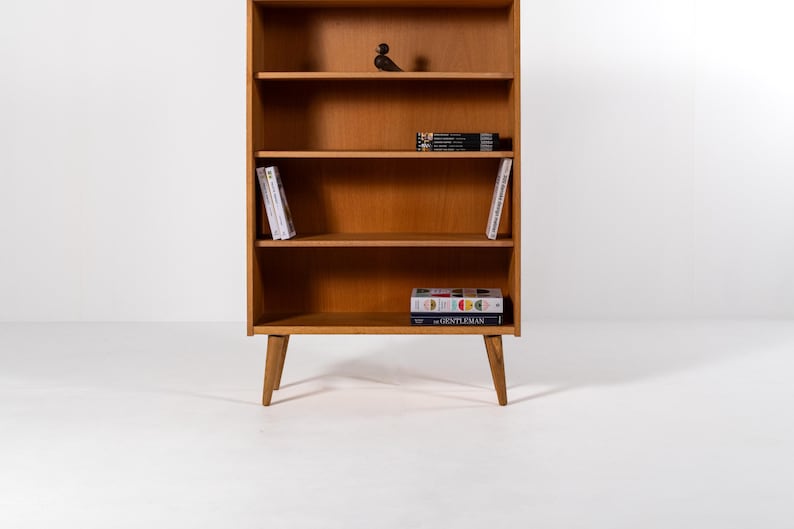 Puede incluir: Una estanter&iacute;a de madera de estilo moderno de mediados de siglo con cuatro estantes y patas anguladas. La estanter&iacute;a est&aacute; llena de libros y un peque&ntilde;o p&aacute;jaro decorativo. La madera tiene un acabado c&aacute;lido y natural.