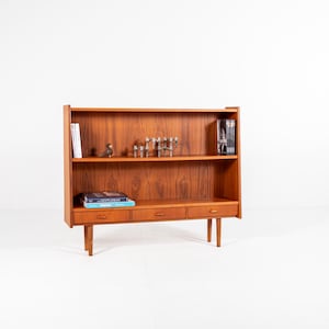 May include: A mid-century modern wooden bookcase with three drawers and two shelves. The bookcase is made of a warm-toned wood and has a minimalist design. Books and decorative items are displayed on the shelves.