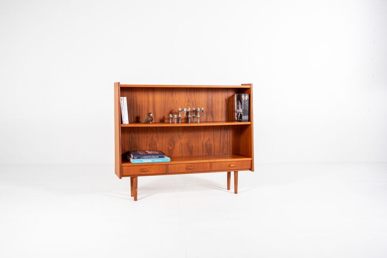 May include: A mid-century modern wooden bookcase with three drawers and two shelves. The bookcase is made of a warm-toned wood and has a minimalist design. Books and decorative items are displayed on the shelves.