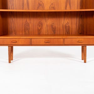 May include: A mid-century modern wooden bookcase with a warm, reddish-brown finish. It features two shelves, three drawers, and four tapered legs. The wood grain is prominently displayed, adding to its vintage appeal.