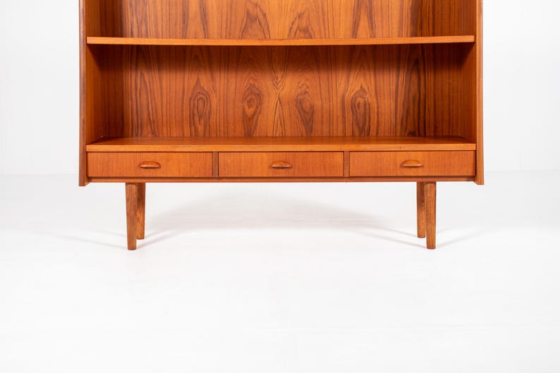 May include: A mid-century modern wooden bookcase with a warm, reddish-brown finish. It features two shelves, three drawers, and four tapered legs. The wood grain is prominently displayed, adding to its vintage appeal.