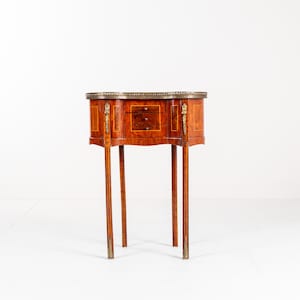 May include: An antique wooden side table with a curved front and three slender legs. The table has two small drawers and decorative brass hardware. The wood is a warm, reddish-brown colour. The table is set against a white background.