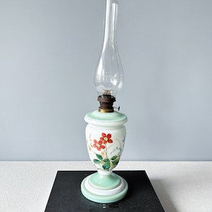 Puede incluir: Una lámpara de aceite vintage con una base de vidrio blanco y verde decorada con diseños florales rojos, verdes y blancos. La lámpara tiene una chimenea de vidrio transparente y un quemador de metal.