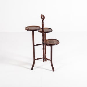 May include: A three-tiered, dark brown wooden plant stand. Each circular tier has a decorative edge. The stand has a handle at the top and is supported by three curved legs. The stand is approximately 91 cm tall.